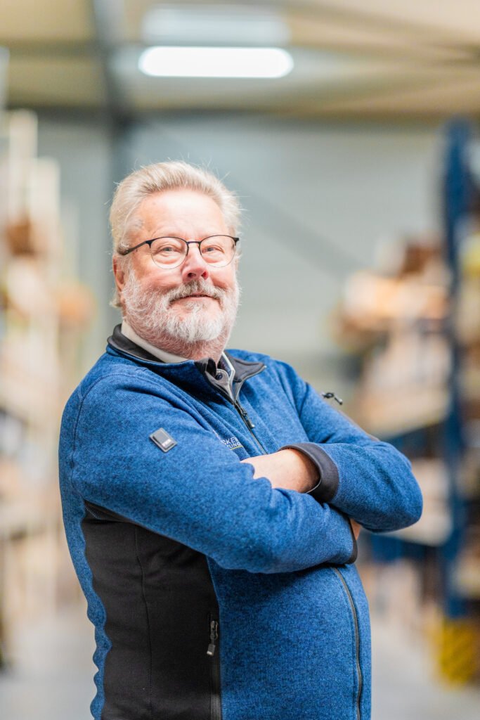 Older man with glasses and a blue jacket stands confidently with arms crossed in a warehouse.