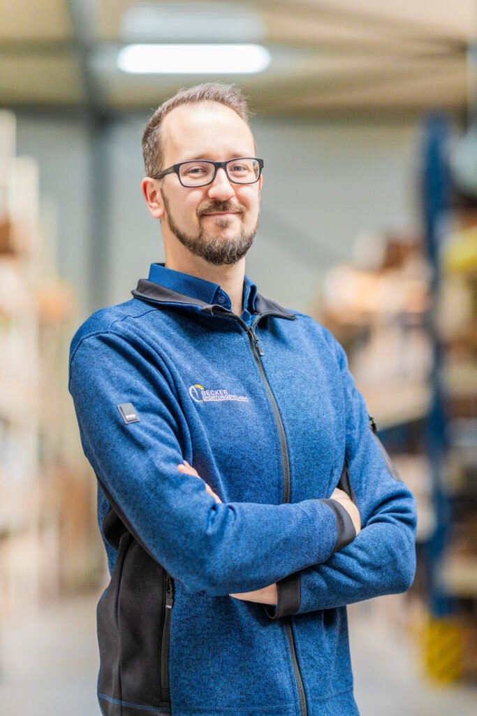 Man with glasses and a beard, wearing a blue jacket, standing in a warehouse with arms crossed, smiling.