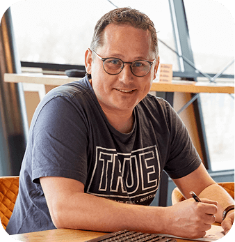 Man with glasses smiling, sitting at a desk with a pen in hand, wearing a "TRUE" t-shirt.