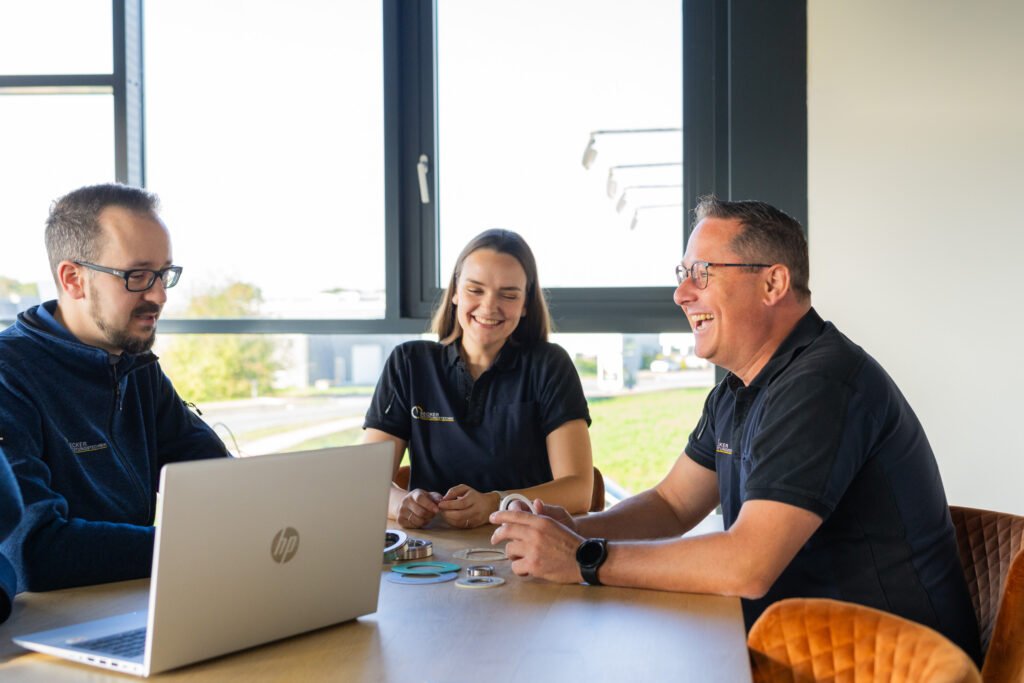 Three people sit at a table with a laptop, smiling and talking in a bright, modern office.