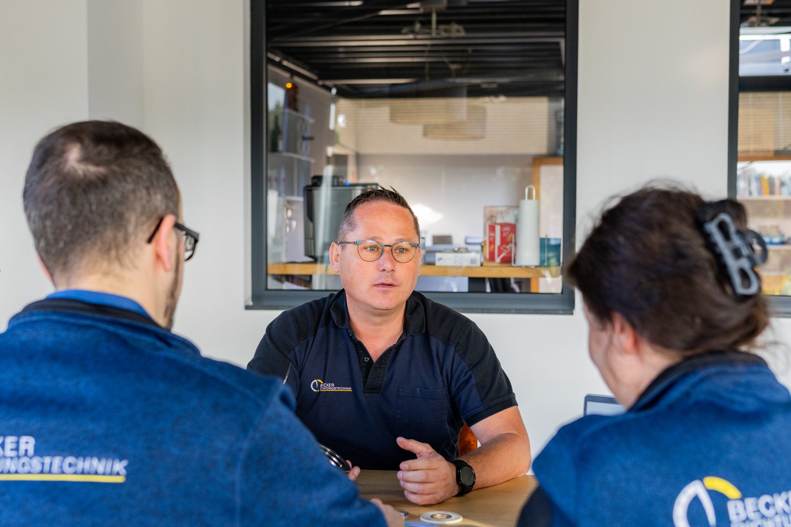 Three people in a meeting, one speaking while the others listen, seated at a table in an office setting.