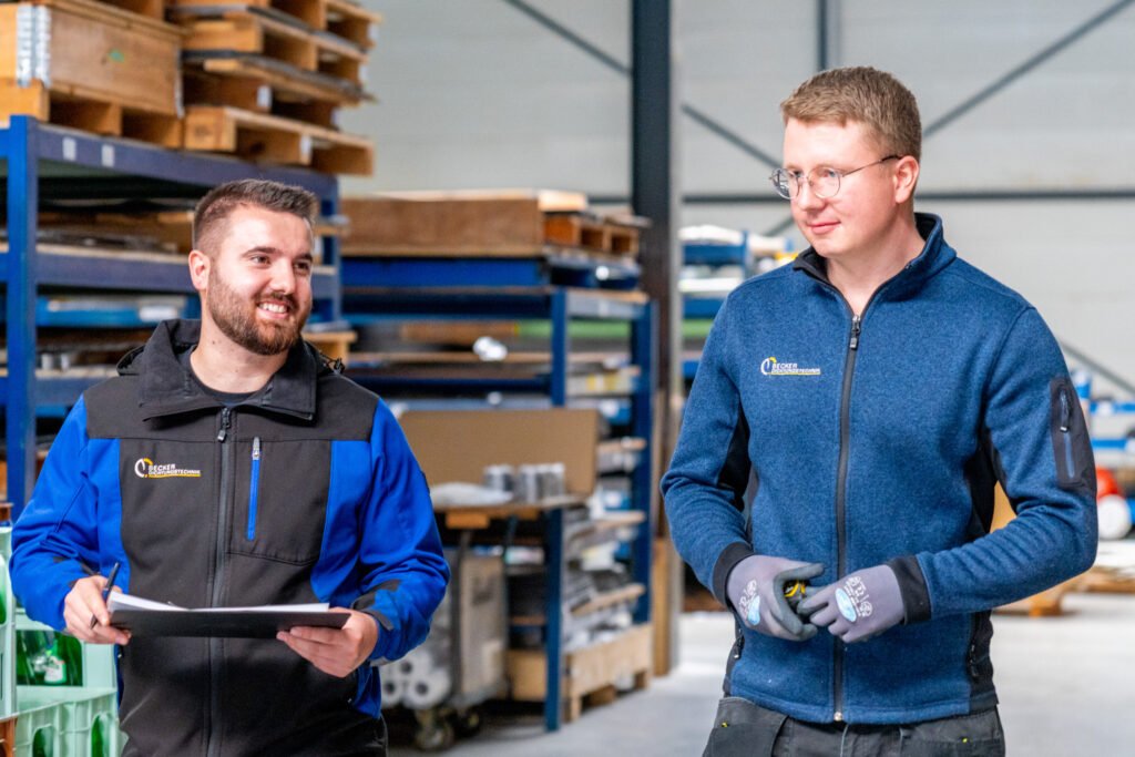 Two men in work uniforms talk and smile in a warehouse with shelves and tools in the background.