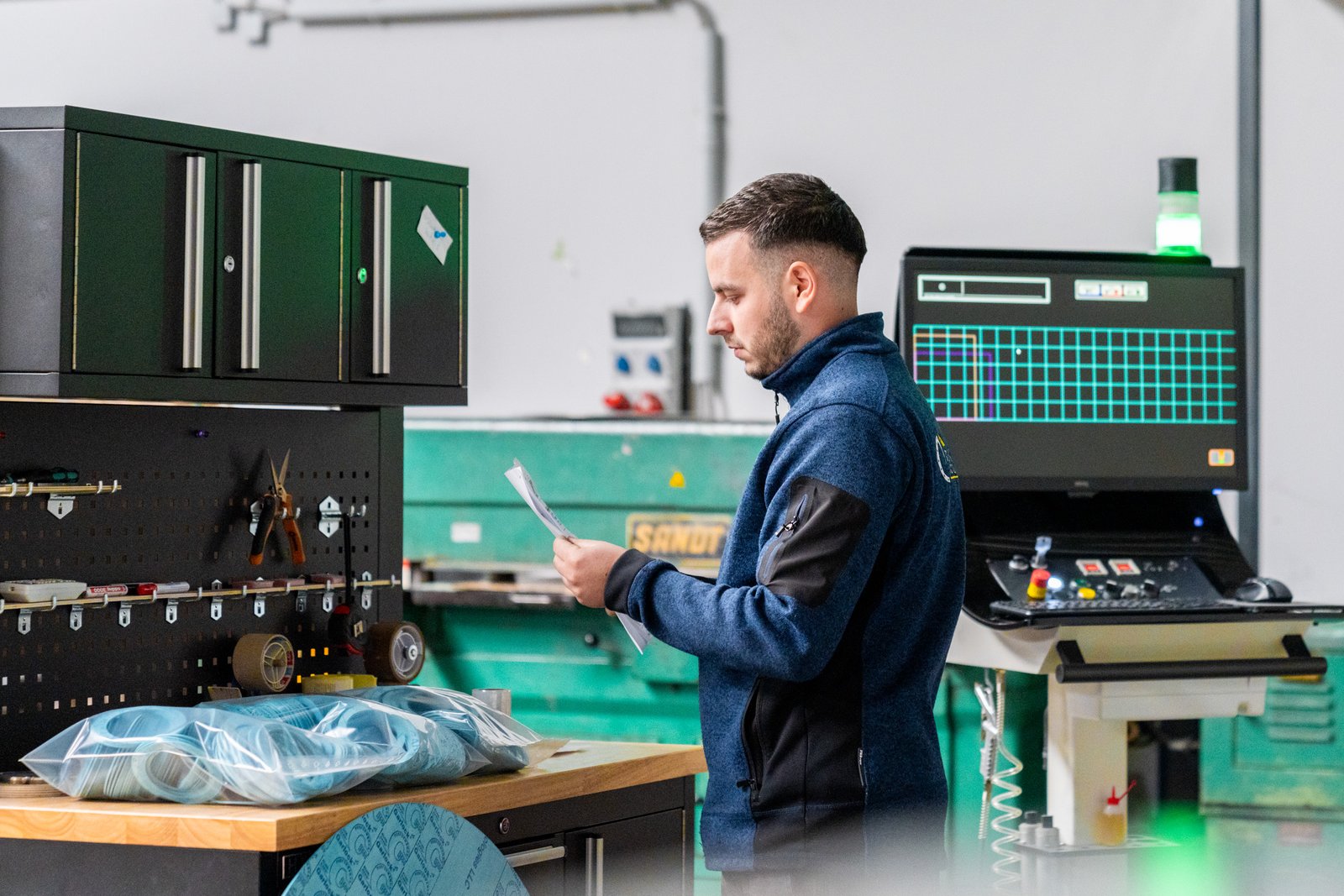 A man reads papers at a workstation in a factory, with equipment and a monitor displaying data nearby.