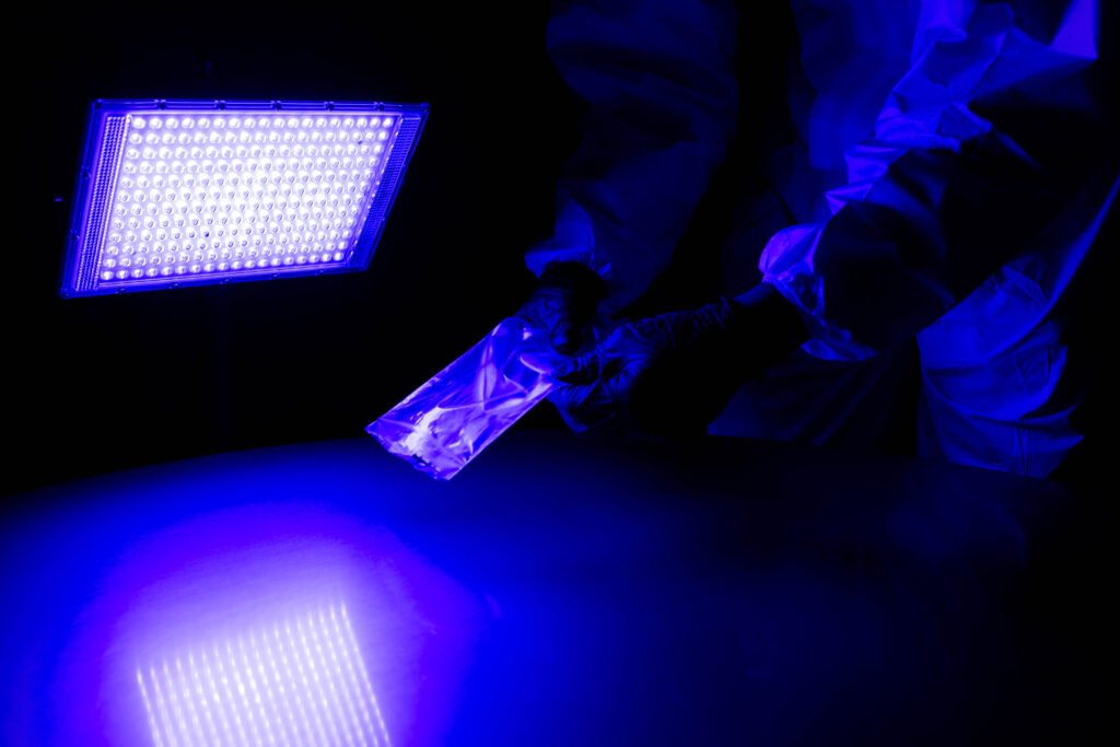 Person in gloves examines evidence under blue forensic light in a dark room.