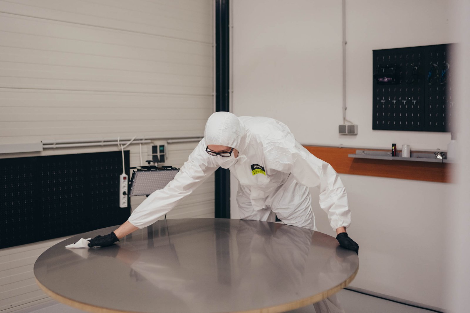 Person in protective suit and gloves cleaning a round metal table in a laboratory or sterile environment.